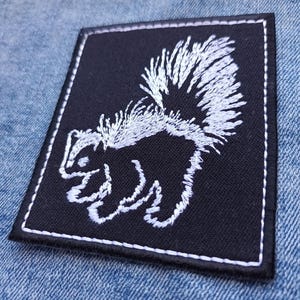 Op de afbeelding: Vierkantige zwarte patch met een wit geborduurd stinkdierontwerp. Het stinkdier wordt in profiel weergegeven, met zijn staart prominent aanwezig. De patch heeft een witte gestikte rand, op een blauwe denim achtergrond.