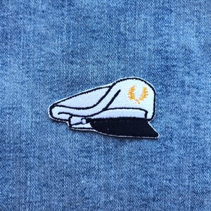 May include: Embroidered patch featuring a white captain's hat with a black peak and a gold laurel wreath. The patch is set against a blue denim fabric.
