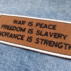 May include: A rectangular brown patch with the text "WAR IS PEACE, FREEDOM IS SLAVERY, IGNORANCE IS STRENGTH" in black. The patch has a white border and is set against a blue denim background.