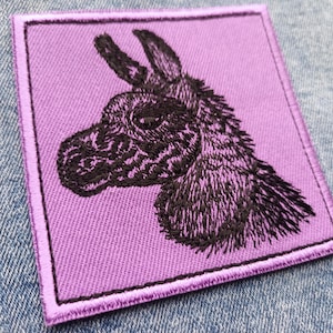 May include: A square embroidered patch with a black donkey head on a lavender background. The patch has a lavender border. The donkey's fur is intricately detailed. The patch is designed to be sewn onto clothing or accessories.