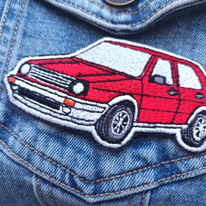 Pode incluir: Patch bordado com um carro vermelho com teto branco e pneus pretos. O patch do carro tem uma borda branca e é colocado sobre um fundo de ganga azul.