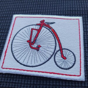 Könnte beinhalten: Gestickter Aufnäher mit einem roten Hochrad-Fahrrad-Design auf weißem Hintergrund, umrahmt von einem roten Rand. Der Aufnäher ist rechteckig und auf einem strukturierten dunkelblauen Stoff angebracht.
