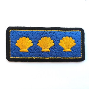 May include: Embroidered patch with three gold scallop shell designs on a blue background. The patch has a black border with yellow stitching, creating a decorative and nautical-themed accessory.