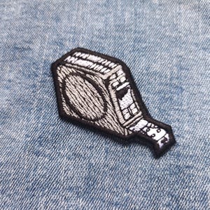 May include: A black and white embroidered patch featuring a vintage boombox design. The patch has a detailed outline and a textured fill, with a black border. The boombox design includes a speaker, buttons, and a handle.
