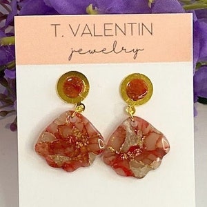 Op de afbeelding: Gouden oorbellen met rode en gouden marmeren harsbloemenhangers. De oorbellen bevinden zich op een witte kaart met de tekst "T. VALENTIN jewelry" en "www.tvalentinjewelry.etsy.com".