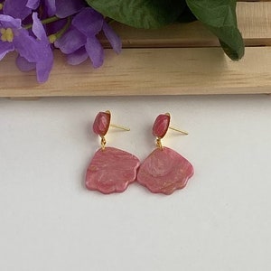 May include: A pair of pink and gold shell-shaped earrings with a marbled effect. The earrings are dangle style with a small gold bead at the top.