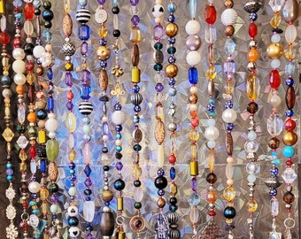 May include: A colorful beaded curtain with strands of various colors, including blue, green, red, and gold. The beads are made of glass, wood, and metal. The curtain is hanging from a metal rod.