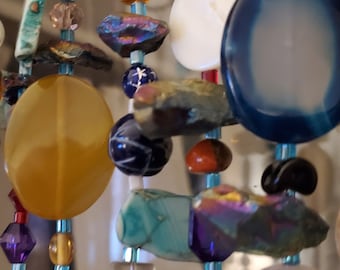 Op de afbeelding: Een kleurrijke kralenketting met verschillende vormen en maten kralen, waaronder ronde, ovale en onregelmatige vormen. De kralen zijn gemaakt van verschillende materialen, zoals glas, metaal en steen. De ketting is geregen op een doorzichtig koord en heeft een verscheidenheid aan kleuren, waaronder blauw, groen, geel, rood en paars.
