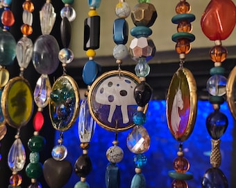 May include: A colorful beaded curtain with various charms, including a mushroom, a unicorn, and a sun with a star. The beads are made of glass, metal, and wood.