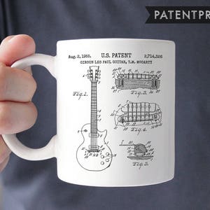 Pode incluir: Caneca de cerâmica branca com uma imagem em preto e branco de um desenho de patente de uma guitarra Gibson Les Paul. O número da patente é 2.714.326 e a data é 2 de agosto de 1955. O texto "U.S. PATENT" e "GIBSON LES PAUL GUITAR, T.M. MOCARTY" também estão incluídos na imagem.