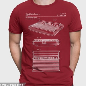 Op de afbeelding: Een rood T-shirt met een witte print van een patenttekening voor een gameconsole. De tekening toont de console vanuit verschillende hoeken. De tekst op het T-shirt luidt "United States Patent", "Hardy et al.", "GAME CONTROL CONSOLE", "FIG 1", "FIG 2", "FIG 4", "Des. 251,143" en "Feb. 20, 1979".