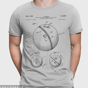 Pode incluir: Uma camiseta cinza com uma ilustra&ccedil;&atilde;o em preto e branco de um desenho de patente de bola de boliche. O n&uacute;mero da patente &eacute; 3.318.598 e o inventor &eacute; H. Ruskin. O texto "Bowling Ball Having Means for Indicating a Correct Hooked Delivery" tamb&eacute;m est&aacute; inclu&iacute;do.