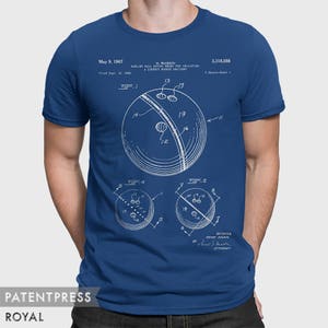 Pode incluir: Uma camiseta azul com uma estampa branca de um desenho de patente de bola de boliche. O n&uacute;mero da patente &eacute; 3.318.598 e o inventor &eacute; H. Ruskin. O texto na camiseta diz "Bowling Ball Having Means For Indicating A Correct Hooked Delivery".