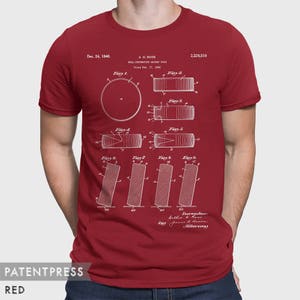 May include: A red t-shirt with a white print of a patent drawing for a "Roll-Preventing Hockey Puck" by A.H. Rogs, filed February 17, 1940. The patent number is 2,226,516. The drawing shows different views of the puck, with the text "Dec. 24, 1940" and "Inventor: Arthur H. Rogs, by James R. Howard, Attorney" at the bottom.
