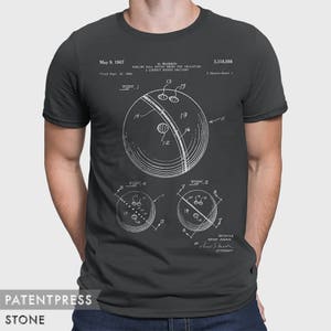 Pode incluir: Uma camiseta cinza escura com uma estampa branca de um desenho de patente de bola de boliche. O n&uacute;mero da patente &eacute; 3.318.598 e a data da patente &eacute; 9 de maio de 1967. O inventor &eacute; H. Ruskin.