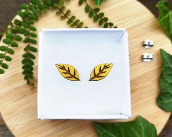 Puede incluir: Un par de pendientes en forma de hoja amarilla en una caja blanca. Los pendientes están hechos de madera y tienen un patrón de vetas marrones oscuras.