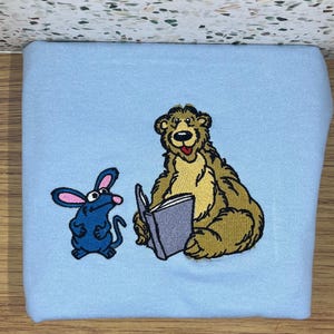 May include: A light blue sweatshirt featuring an embroidered design of a cartoon bear reading a book, with a blue mouse looking on. The bear is brown and tan, and the mouse is blue with pink ears. The book is gray.