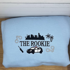 May include: Light blue sweatshirt with an embroidered design featuring the text "THE ROOKIE", a police car, handcuffs, a cityscape, and palm trees. The design is in black and white.