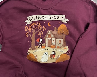 May include: Burgundy sweatshirt with a graphic featuring a house, trees, pumpkins, and the text "Gilmore Ghouls Where You Haunt, I Will Follow." The design has a Halloween theme with a ghost and figures.