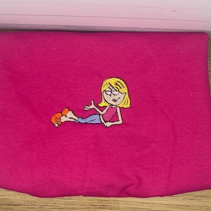 May include: A folded magenta fabric with an embroidered cartoon character. The character has blonde hair, glasses, a pink top, blue pants, and orange shoes. The embroidery is detailed and colorful, set against the solid magenta background.