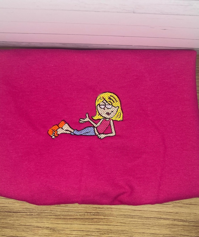 May include: A folded magenta fabric with an embroidered cartoon character. The character has blonde hair, glasses, a pink top, blue pants, and orange shoes. The embroidery is detailed and colorful, set against the solid magenta background.