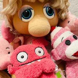 May include: A collection of plush toys, including a large pig-like character with blonde hair and a lace headpiece, surrounded by smaller pink stuffed animals. The toys have large eyes and are in various shades of pink.