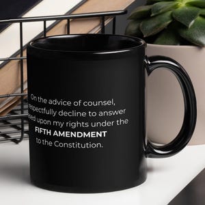 May include: A black ceramic mug featuring white printed text that reads: On the advice of counsel, I respectfully decline to answer based upon my rights under the FIFTH AMENDMENT to the Constitution. The mug has a standard handle and cylindrical body.