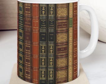 May include: White ceramic coffee mug featuring a repeating pattern of antique book spines in shades of brown, red, and green. The mug has a curved handle and sits on a beige coaster. The design evokes a vintage library aesthetic.