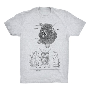 Könnte beinhalten: Ein graues T-Shirt mit einer Schwarzweißillustration einer Furby-Spielzeugpatentzeichnung. Die Zeichnung zeigt die inneren Komponenten des Spielzeugs und das äußere Design des Spielzeugs. Der Text "FIG. 8A" und "FIG. 11" ist in der Zeichnung enthalten.