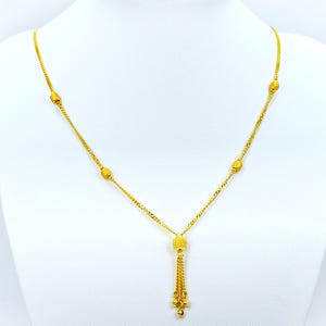 May include: A gold necklace with a tassel pendant and small round beads. The necklace is on a white background.