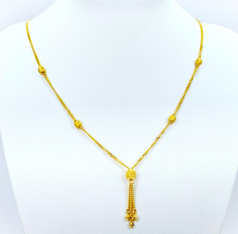 May include: A gold necklace with a tassel pendant and small round beads. The necklace is on a white background.