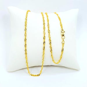 May include: A gold-colored necklace with a twisted rope design, resting on a white pillow. The necklace features a delicate chain with a secure clasp. The twisted pattern catches the light, creating a shimmering effect. The jewelry is displayed against a plain background.