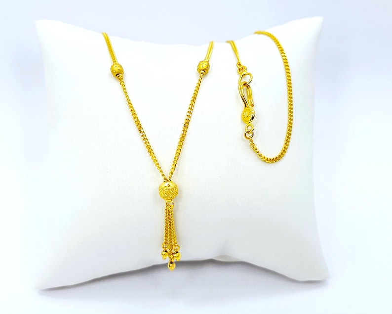 May include: A gold chain necklace with a tassel pendant. The chain has small round beads and the pendant has a larger round bead with a textured surface.