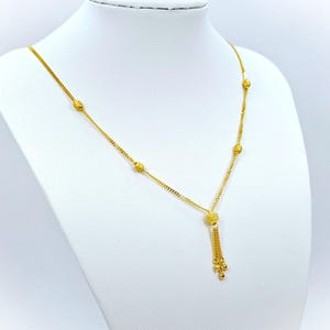 May include: A gold necklace with a tassel pendant and small round beads. The necklace is on a white mannequin.