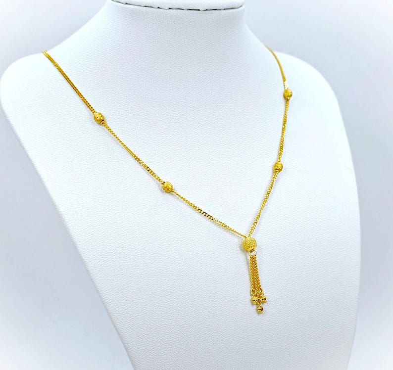 May include: A gold necklace with a tassel pendant and small round beads. The necklace is on a white mannequin.