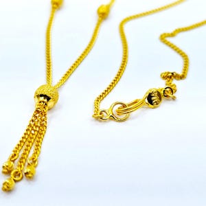 May include: A gold chain necklace with a tassel pendant. The pendant is a small, round ball with a textured surface. The chain is a delicate, curb chain.