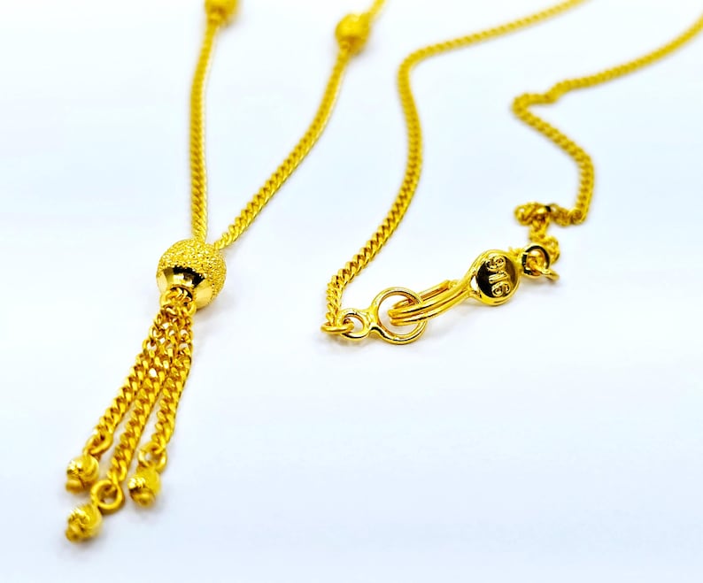 May include: A gold chain necklace with a tassel pendant. The pendant is a small, round ball with a textured surface. The chain is a delicate, curb chain.