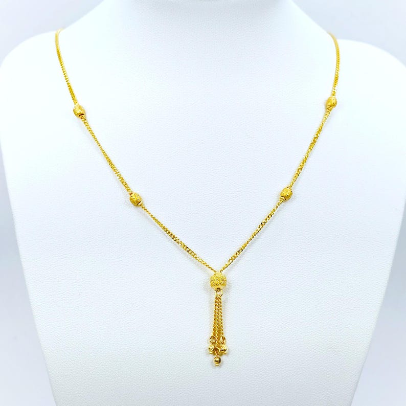 May include: A gold necklace with a tassel pendant and small round beads. The necklace is on a white background.