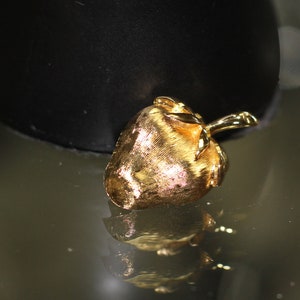 Puede incluir: Un colgante de color dorado en forma de corazón con un pequeño tallo. El colgante está sobre una superficie oscura.