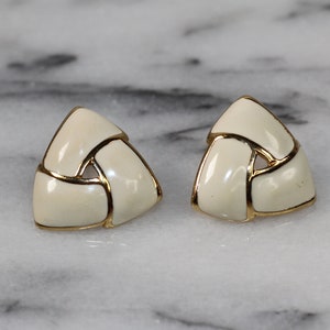 May include: A pair of gold tone earrings with a white enamel design. The earrings are shaped like a triangle and have a woven pattern.