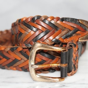 K&ouml;nnte beinhalten: Ein geflochtener Lederg&uuml;rtel mit einer silbernen Schnalle. Der G&uuml;rtel ist aus braunem, schwarzem und braunem Leder.