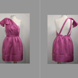 Op de afbeelding: Een roze jurk met één schouder en een strikdetail op de band. De jurk heeft een aansluitend bovenlijf en een volle rok.