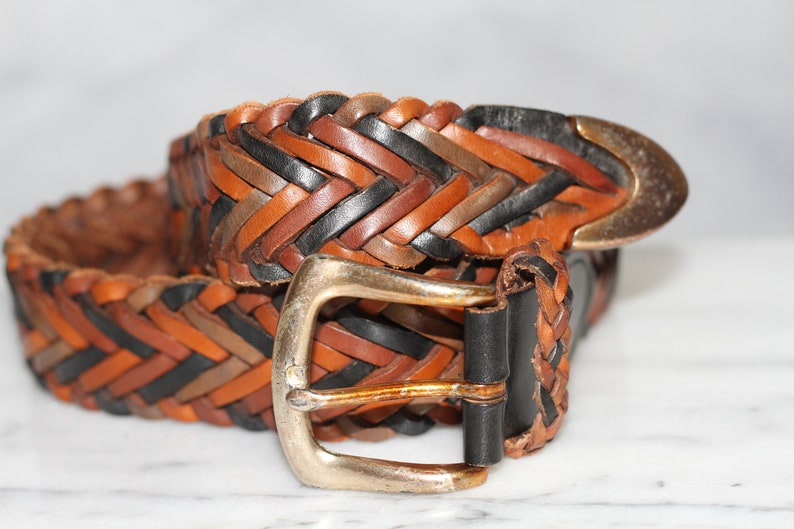 K&ouml;nnte beinhalten: Ein geflochtener Lederg&uuml;rtel mit einer silbernen Schnalle. Der G&uuml;rtel ist aus braunem, schwarzem und braunem Leder.