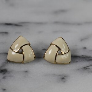 May include: A pair of gold tone earrings with a white enamel design. The earrings are shaped like triangles and have a woven pattern.