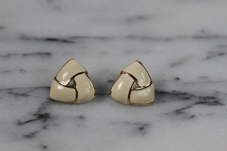 May include: A pair of gold tone earrings with a white enamel design. The earrings are shaped like triangles and have a woven pattern.