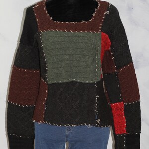 K&ouml;nnte beinhalten: Ein Patchwork-Pullover mit braunen, gr&uuml;nen, schwarzen und roten Strickpanels. Der Pullover hat einen Rundhalsausschnitt und lange &Auml;rmel.