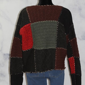 K&ouml;nnte beinhalten: Ein schwarzer, roter und gr&uuml;ner Patchwork-Pullover mit Zopfmuster. Der Pullover hat einen Rundhalsausschnitt und lange &Auml;rmel.