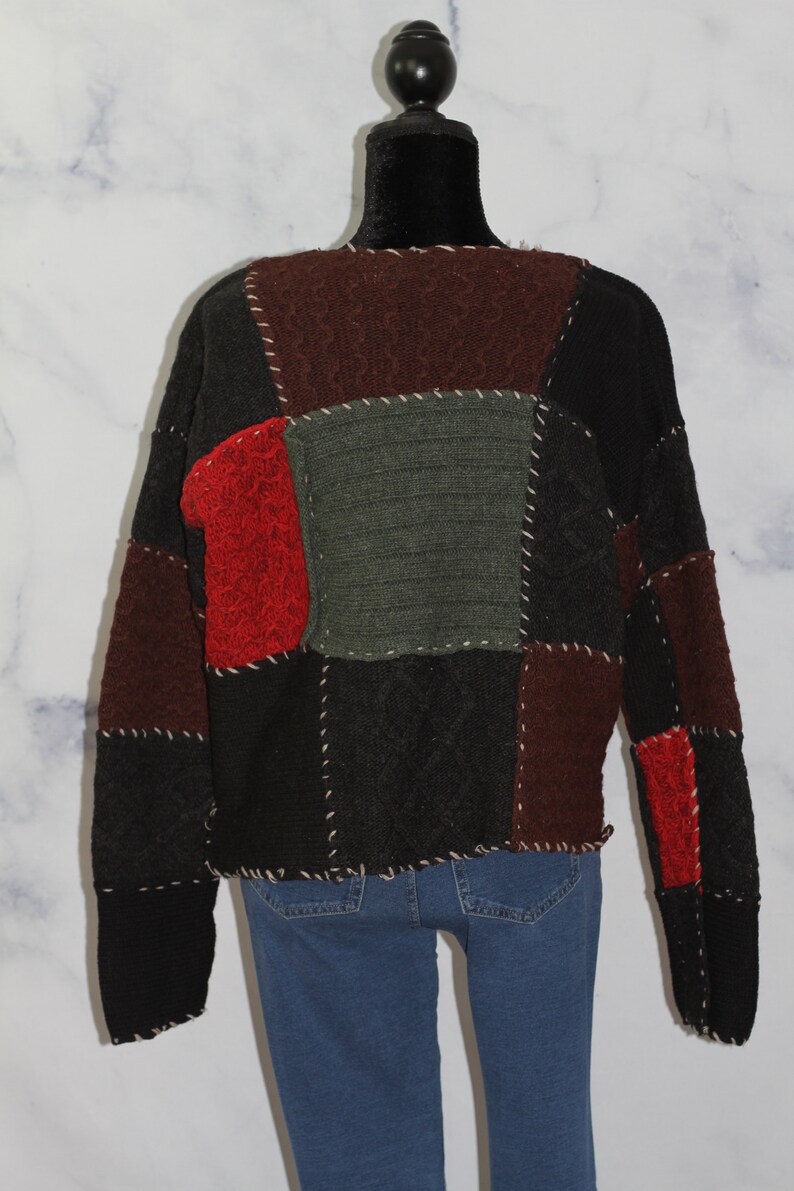 K&ouml;nnte beinhalten: Ein schwarzer, roter und gr&uuml;ner Patchwork-Pullover mit Zopfmuster. Der Pullover hat einen Rundhalsausschnitt und lange &Auml;rmel.