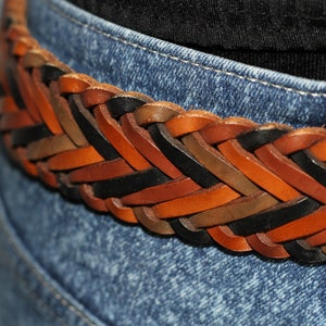 K&ouml;nnte beinhalten: Ein geflochtener Lederg&uuml;rtel in Braun, Schwarz und Tan. Der G&uuml;rtel ist im Fischgr&auml;tenmuster gewebt und wird auf einer blauen Jeans gezeigt.