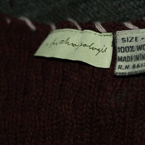 K&ouml;nnte beinhalten: Ein gr&uuml;nes Etikett mit dem Text "anthropologie" und ein braunes Etikett mit dem Text "SIZE - L 100% WOOL MADE IN INDIA R.N. 66170" befestigt an einem dunkelbraunen Strickstoff.